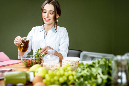 Woman dietitian working on a healthy diet pouring olive oil into the salad sitting at the table full of various healthy food indoorsの写真素材