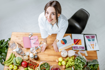 Portrait of a young woman nutritionist in medical uniform standing near the table full of various healthy products indoorsの写真素材