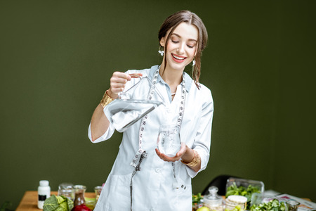 Portrait of a young woman nutritionist in medical gown standing with water in the office with healthy food on the backgroundの写真素材