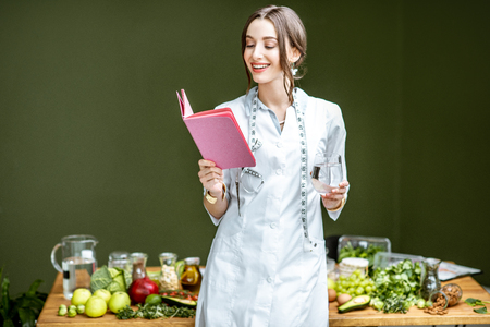 Portrait of a young woman nutritionist in medical gown standing with notebook and glass of water in the office with healthy food on the backgroundの写真素材