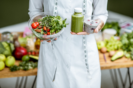 Nutritionist in medical gown holding salad and smoothie drink with healthy food on the background, close-up view with no faceの写真素材