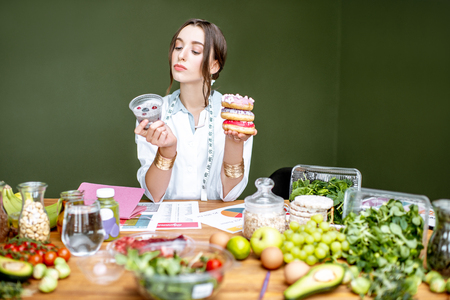 Young woman nutritionist looking on the chia pudding choosing between healthy food and sweet dessert sitting at the table full of different productsの写真素材