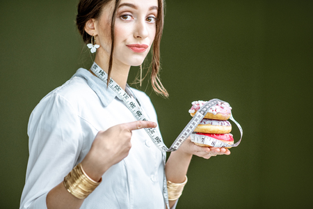 Portrait of a sad woman nutritionist in medical gown with donuts and tape measure on the green background. Unhealthy eating and adiposity conceptの写真素材