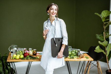 Portrait of a young woman nutritionist in medical gown standing with weights and tape measure in the office with healthy food on the backgroundの写真素材