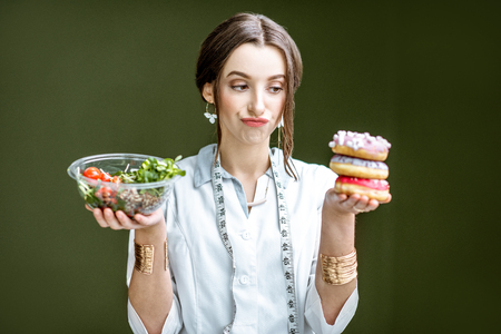 Young woman nutritionist looking on the donuts with sad emotions choosing between salad and unhealthy dessert on the green backgroundの写真素材