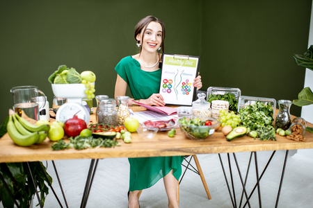 Portrait of a beautiful woman as a dietitian showing drawings on the topic of healthy food sitting with various healthy food ingredients indoorsの写真素材