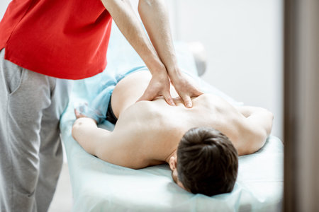 Young male masseur doing back massage to a man in the white cabinet of rehabilitation clinic