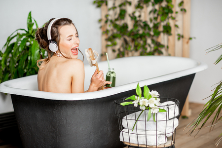 Young relaxed woman listening to the music and drinking smoothie while lying in the retro bathtub at the beautiful bathroom with green plantsの写真素材