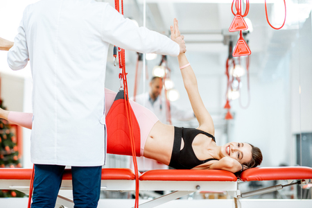 Senior physiotherapist doing treatment to a young woman with spine problems with suspension medical equipment at the rehabilitation clinicの写真素材