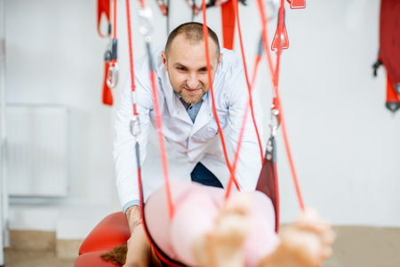 Senior physiotherapist doing treatment to a young woman with spine problems with suspension medical...
