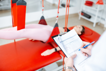 Physiotherapist writing medical results during the suspension treatment for the young woman with spine problemsの写真素材