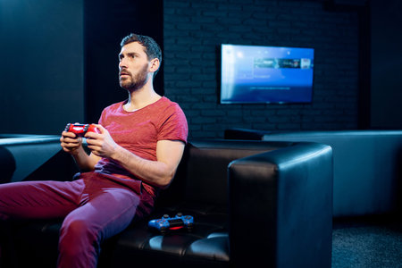 Man playing video games with gaming console and joy stick sitting on the couch in the dark room of...