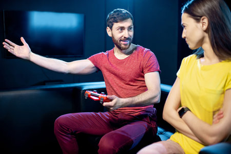 Woman offended by a boyfriend playing video games with gaming console sitting on the couch in the...