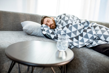 Man feeling sick lying on the couch covered with blanket with pills and glass of water on the table...