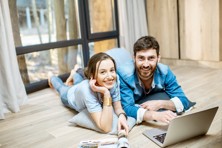 Portrait of a happy couple lying on the floor with laptop and magazines, relaxing at the cozy homeの写真素材