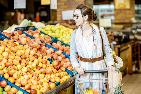 Young woman buying food, standing with shopping cart full of products in the supermarket with fruits on the backgroundの写真素材