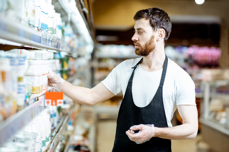 Shop worker looking on the bottles with milk standing near the shelves with dairy products in the supermarketの写真素材