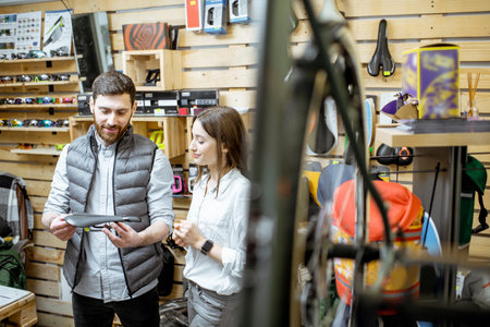 Salesman helping young woman customer to choose paddles standing near the shelves with bicyles parts in the sports shop