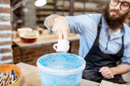 Handsome worker painting clay jugs, diving products into the bucket with blue paint at the pottery...