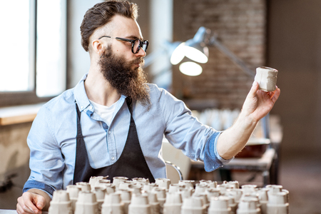 Handsome bearded man working with ceramic workpieces at the working place in the pottery shopの写真素材