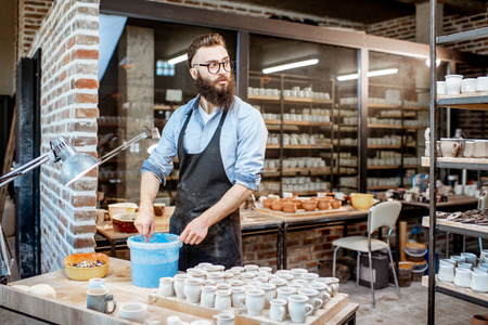 Handsome worker painting clay jugs, diving products in the bucket with blue paint at the pottery shopの写真素材