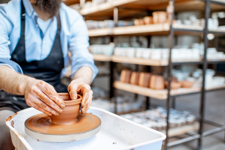 Man making clay jug forming shape by hands on the pottery wheel indoors, close-up viewの写真素材