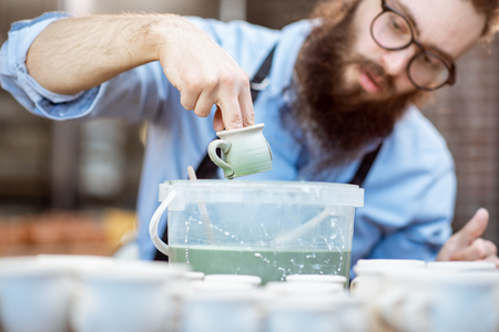 Handsome creative bearded worker painting clay jugs, diving products into the bucket with green paint at the pottery shopの写真素材
