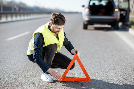 Man in road vest putting emergency triangle sign on the highway with broken car on the background