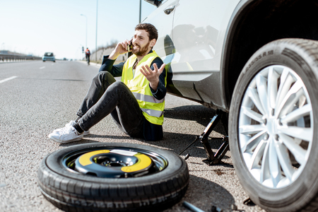 Man calling the road assistance sitting near the car with broken wheel on the roadsideの写真素材