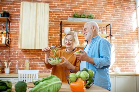 Cheerful senior couple eating salad standing together with healthy food on the kitchen at home. Concept of healthy nutrition in older ageの写真素材
