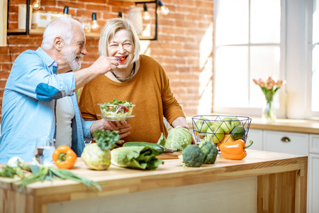 Cheerful senior couple eating salad standing together with healthy food on the kitchen at home. Concept of healthy nutrition in older ageの写真素材
