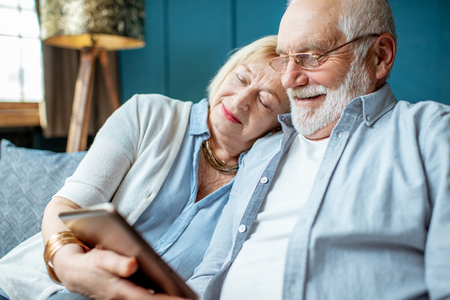 Lovely senior couple dressed casually using digital tablet while sitting together on the comfortable couch at homeの写真素材