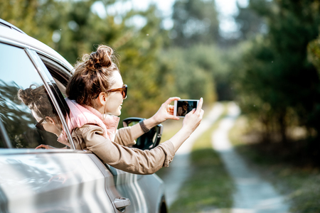 Young woman enjoying the trip, photographing out the window with phone on a picturesque road in the woodsの写真素材