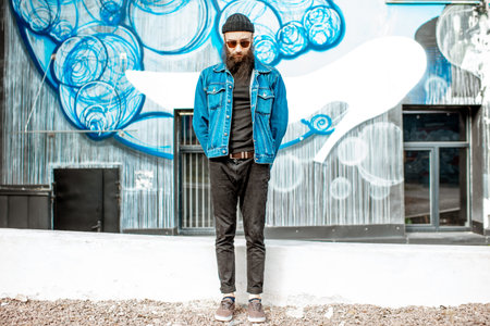Portrait of a stylish bearded man dressed in hat and jacket standing outdoors on the colorful wall...