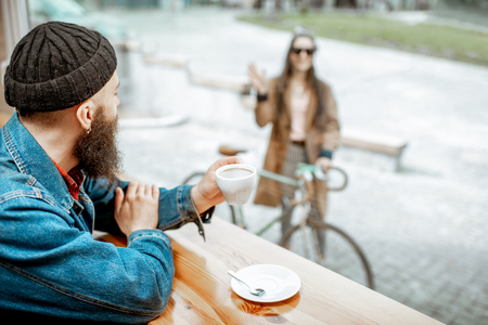 Stylish man meeting his girlfriend with bicycle while sitting with coffee near the window at the cafeの写真素材