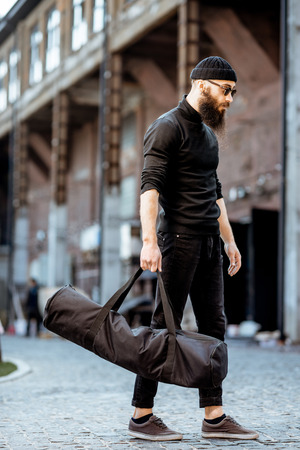 Portrait of a serious bearded man as a killer dressed in black tight clothes holding bag with weapons outdoors on the industrial backgroundの写真素材