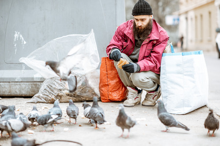 Portrait of a homeless bearded beggar feeding pigeons near the trash in the city. Concept of poverty and unemploymentの写真素材