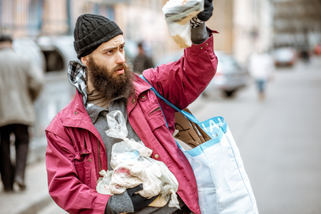Portrait of a homeless bearded beggar standing with bag and some finded food near the trash containers in the city. Concept of povertyの写真素材