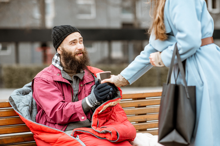 Woman helping homeless beggar giving some hot drink outdoors. Concept of helping poor peopleの写真素材