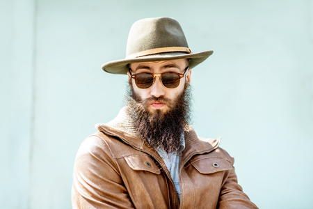 Portrait of a stylish bearded man dressed in jacket and hat on the light turquoise background outdoorsの写真素材