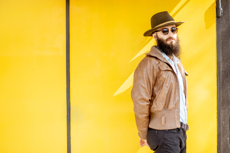 Portrait of a stylish bearded man dressed in jacket and hat on the bright yellow background outdoorsの写真素材