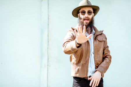 Portrait of a stylish bearded man showing stop with hand on the light turquoise background outdoorsの写真素材