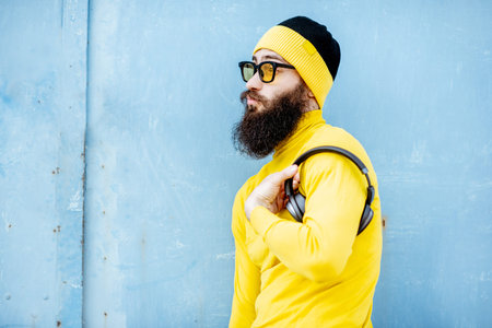 Portrait of a stylish bearded man in yellow clothes enjoying music with headphones on the blue...