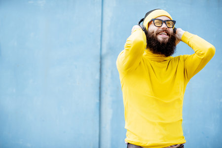 Portrait of a stylish bearded man in yellow clothes enjoying music with headphones on the blue backgroundの写真素材