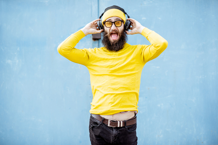 Portrait of a stylish bearded man in yellow clothes enjoying music with headphones on the blue backgroundの写真素材
