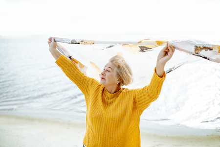 Senior woman in bright sweater enjoying sea breeze, holding scarf above the head on the sandy beachの写真素材