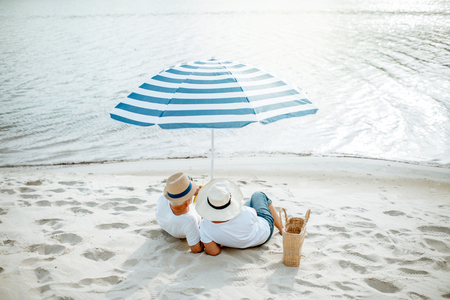 Senior couple sitting together under umbrella on the sandy beach, enjoying their retirement near the sea, rear viewの写真素材