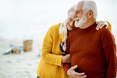 Portrait of a happy senior couple dressed in colorful sweaters hugging on the sandy beach, enjoying free time during retirement near the seaの写真素材