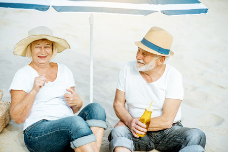Happy senior couple relaxing, lying together with drinks under umbrella on the sandy beach, enjoying their retirement near the seaの写真素材