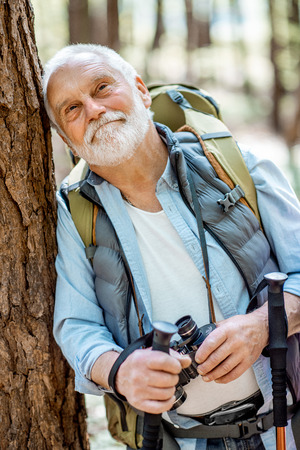 Portrait of a senior man with binoculars and backpack resting near the tree while traveling in the forestの写真素材